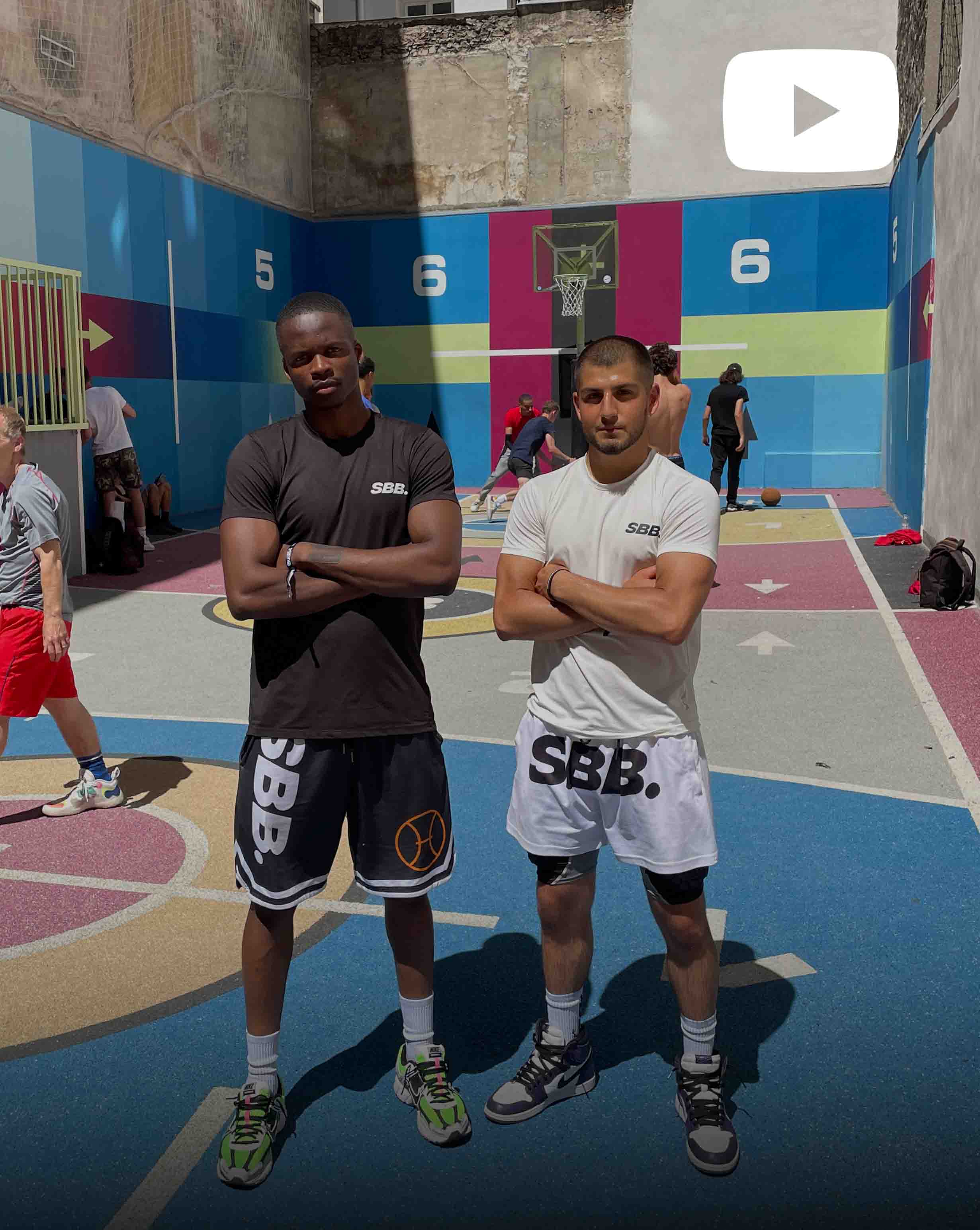Behrad and Denzel from Simply British Ballers (SBB) stand at Pigalle basketball court in Paris, wearing an SBB t-shirt, SBB shorts and SBB socks